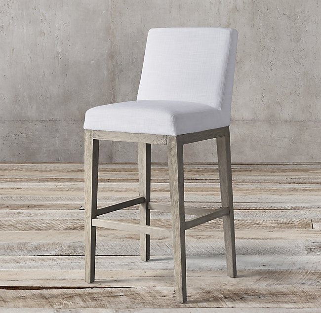 Banqueta Alta Cala tapizada en blanco con patas de madera clara y travesaños apoyapiés