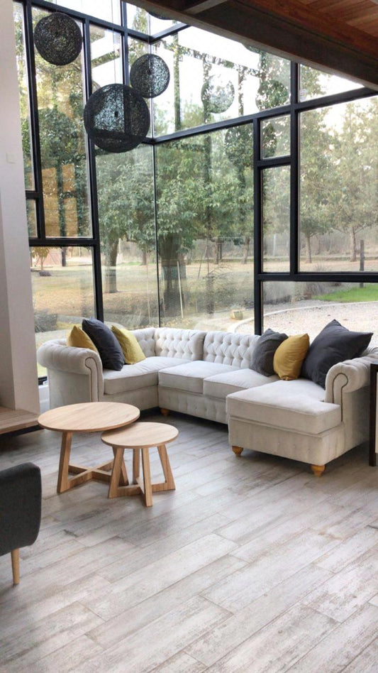 Esquinero Chesterfield beige visto desde otro ángulo, con el chaise, el capitoné completo y las mesas ratonas de madera clara