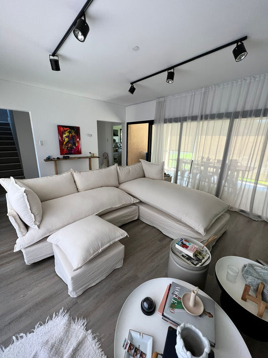 Esquinero Comfy enfundado en tussor crudo con puff al tono, en el living de Maxi con ventanal y cortinas blancas