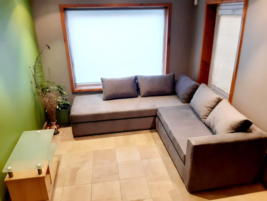Esquinero Cúrcuma gris con base tapizada sin patas a la vista y almohadones de respaldo sueltos, junto a una ventana con marco de madera en el living de Carla