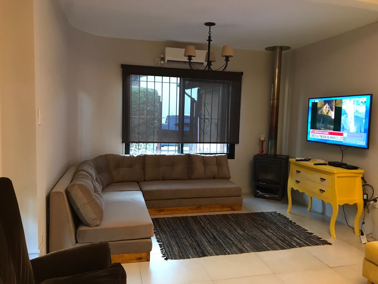 Sillón esquinero amplio de tapizado beige con base de madera, acompañado de cojines mullidos, ubicado en un living con alfombra oscura y ventanal al fondo