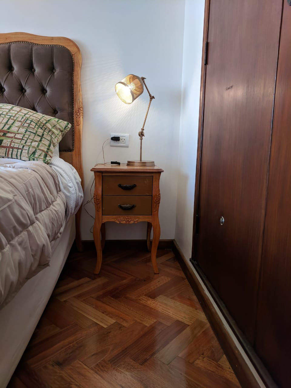 Mesa de Luz Bailarina en madera natural, con dos cajones y patas curvas, junto al respaldo retapizado de Estela