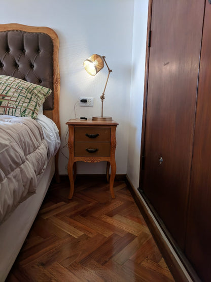 Mesa de Luz Bailarina en madera natural, con dos cajones y patas curvas, junto al respaldo retapizado de Estela