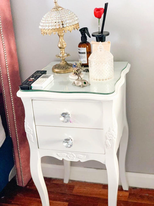 Mesa de Luz Bailarina Valery blanca con tiradores de cristal y un vidrio sobre la tapa, con un velador dorado