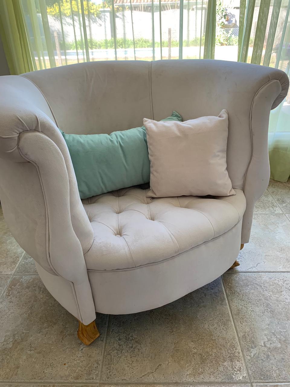 Sillón Brigitte redondo en tapizado beige claro, con capitoné y almohadones verde agua, junto a una ventana con cortinas