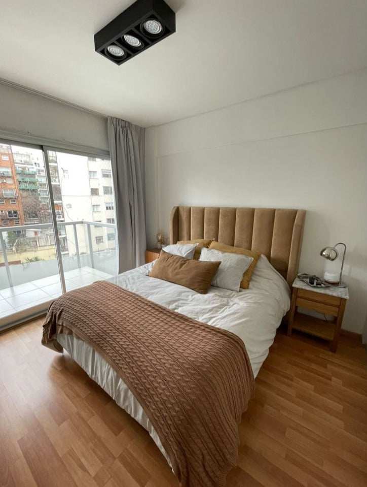 Respaldo Alpes camel de Julieta con mesas de luz de madera y lámpara plateada