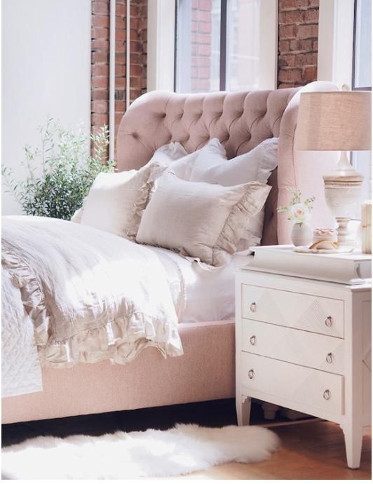 Respaldo de Cama Simona rosa claro con capitoné y borde superior curvo, junto a una mesa de luz blanca
