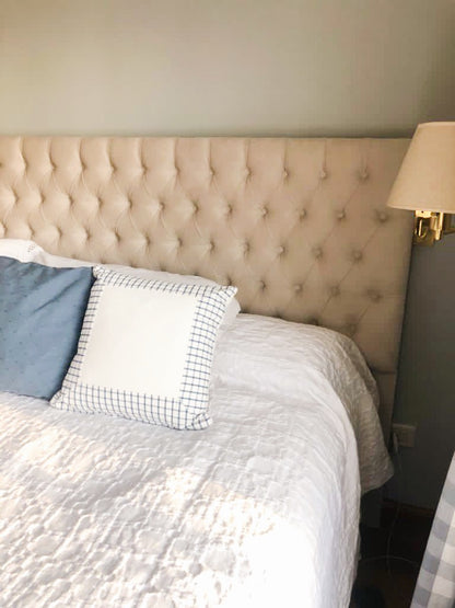 Respaldo de cama amplio y acolchado, tapizado en tela beige clara con un diseño capitoné de botones que forma un patrón de rombos. Su estilo elegante y clásico aporta un toque sofisticado al dormitorio. A ambos lados del respaldo, hay lámparas de pared con pantalla blanca, integradas en repisas flotantes de madera clara, que completan una decoración equilibrada y acogedora.