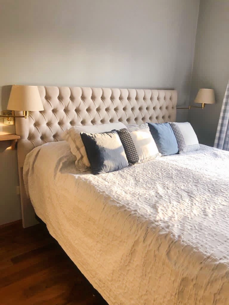 Respaldo de cama amplio y acolchado, tapizado en tela beige clara con un diseño capitoné de botones que forma un patrón de rombos. Su estilo elegante y clásico aporta un toque sofisticado al dormitorio. A ambos lados del respaldo, hay lámparas de pared con pantalla blanca, integradas en repisas flotantes de madera clara, que completan una decoración equilibrada y acogedora.