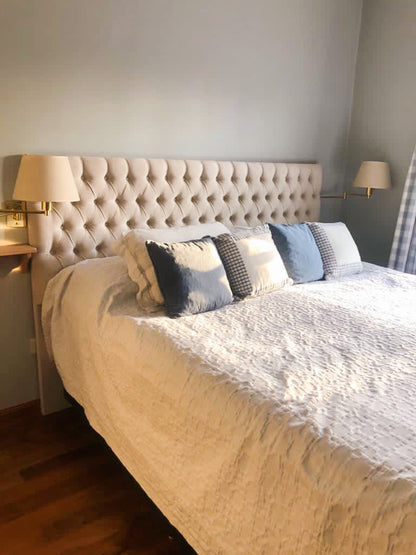 Respaldo de cama amplio y acolchado, tapizado en tela beige clara con un diseño capitoné de botones que forma un patrón de rombos. Su estilo elegante y clásico aporta un toque sofisticado al dormitorio. A ambos lados del respaldo, hay lámparas de pared con pantalla blanca, integradas en repisas flotantes de madera clara, que completan una decoración equilibrada y acogedora.