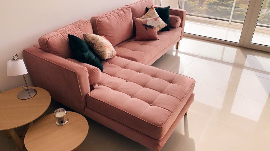 Sillón esquinero Bambú en rosa con asiento de botones y patas de madera, frente al ventanal