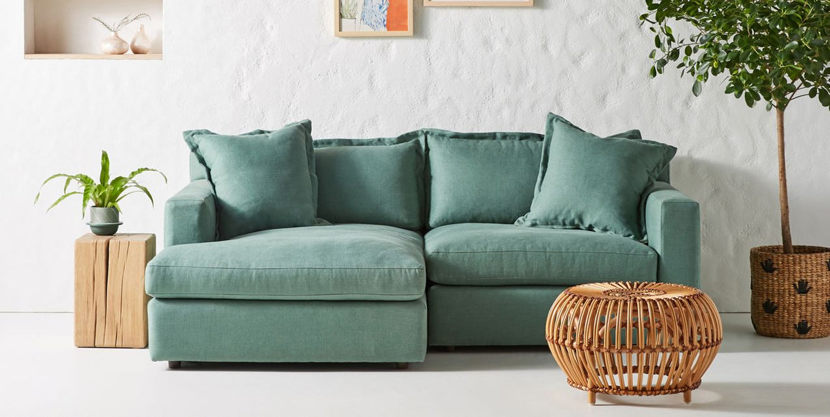 Sillón esquinero Enrique en verde claro junto a una mesa de ratán, con almohadones de respaldo mullidos