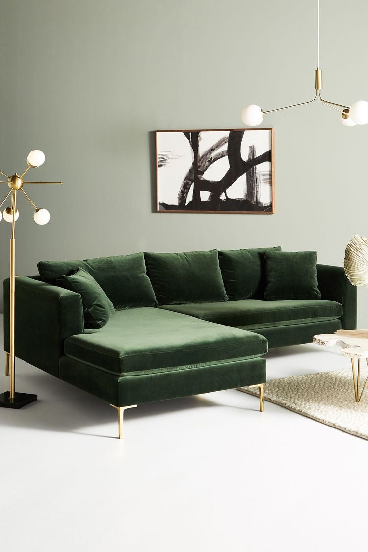 Sillón esquinero Magnolia en pana antimanchas verde inglés con patas doradas, en un living con cuadro y lámpara de pie