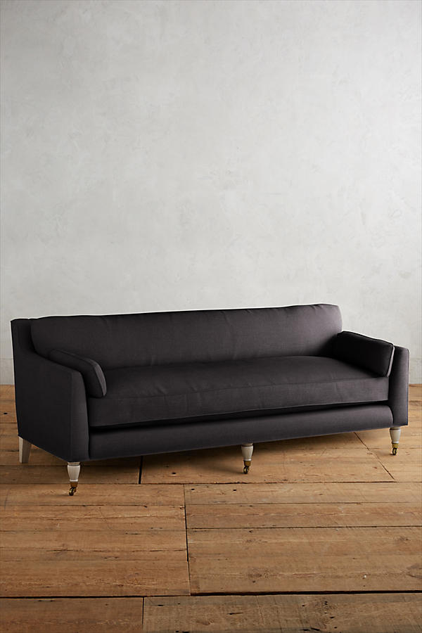 Fotografía de diseño de interiores estilo editorial que muestra un living moderno e industrial sofisticado. En el centro se destaca un sillón de tres cuerpos tapizado en lino gris claro con patas torneadas blancas y rueditas de bronce, idéntico al modelo de referencia. Está ubicado sobre una alfombra de diseño clásico envejecido superpuesta a una gran alfombra de yute