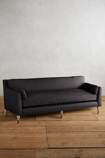 Fotografía de diseño de interiores estilo editorial que muestra un living moderno e industrial sofisticado. En el centro se destaca un sillón de tres cuerpos tapizado en lino gris claro con patas torneadas blancas y rueditas de bronce, idéntico al modelo de referencia. Está ubicado sobre una alfombra de diseño clásico envejecido superpuesta a una gran alfombra de yute