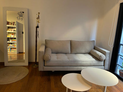 Sofá Bambú en pana gris con asiento capitoné y almohadones cilíndricos, junto a un espejo de pie y una lámpara, en el living de Yamila