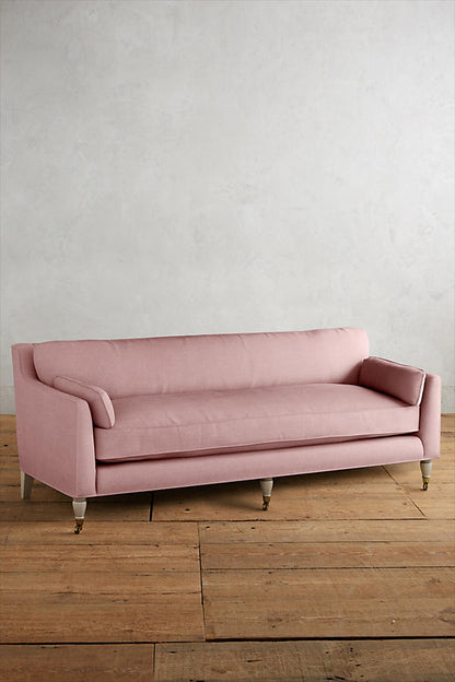 Fotografía de diseño de interiores estilo editorial que muestra un living moderno e industrial sofisticado. En el centro se destaca un sillón de tres cuerpos tapizado en lino gris claro con patas torneadas blancas y rueditas de bronce, idéntico al modelo de referencia. Está ubicado sobre una alfombra de diseño clásico envejecido superpuesta a una gran alfombra de yute